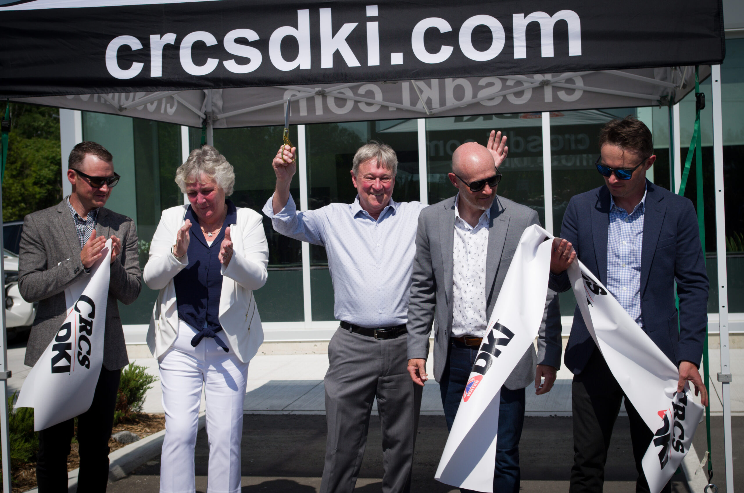Cris raises his arms, scissors still in hand as he celebrates the cutting of the ribbon at the grand opening while Kyle and Robyn clap to his right and Ryan and Matt look at the cut ribbon on Cris's left